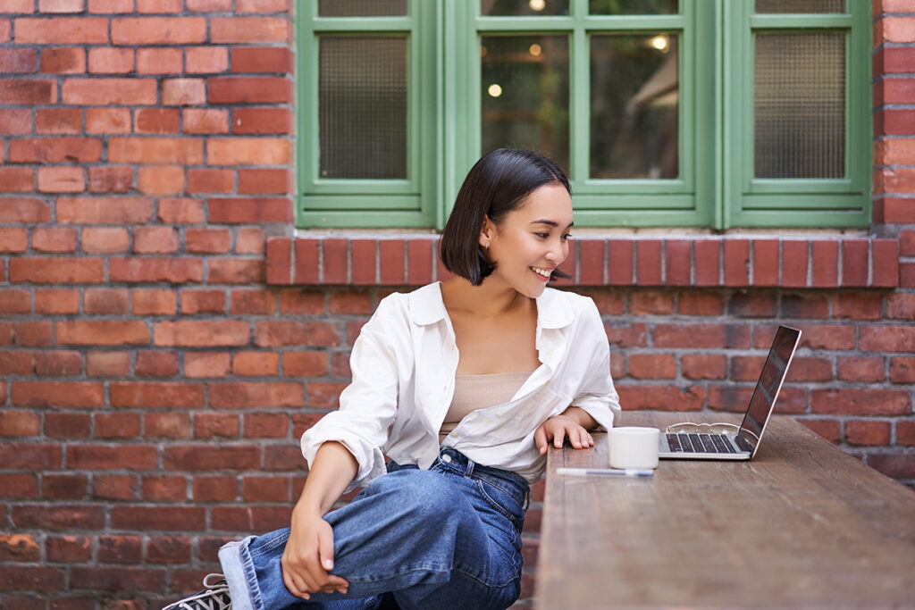 Portrait Of Young Stylish Woman, Influencer Sitting In Cafe With Cup Of Coffee And Laptop, Smiling And Looking Confident